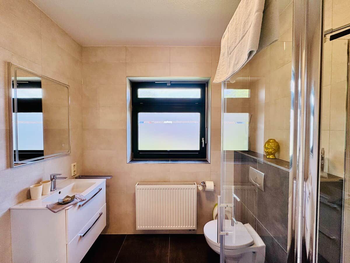 Une salle de bain avec un lavabo blanc, un miroir, une fenêtre, des toilettes et une douche.