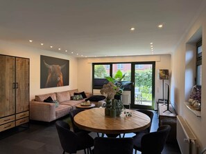 A modern living room with a round wooden table, a sofa, and a large window with a view of greenery. - Margaretha Vacation Home (Varel)
