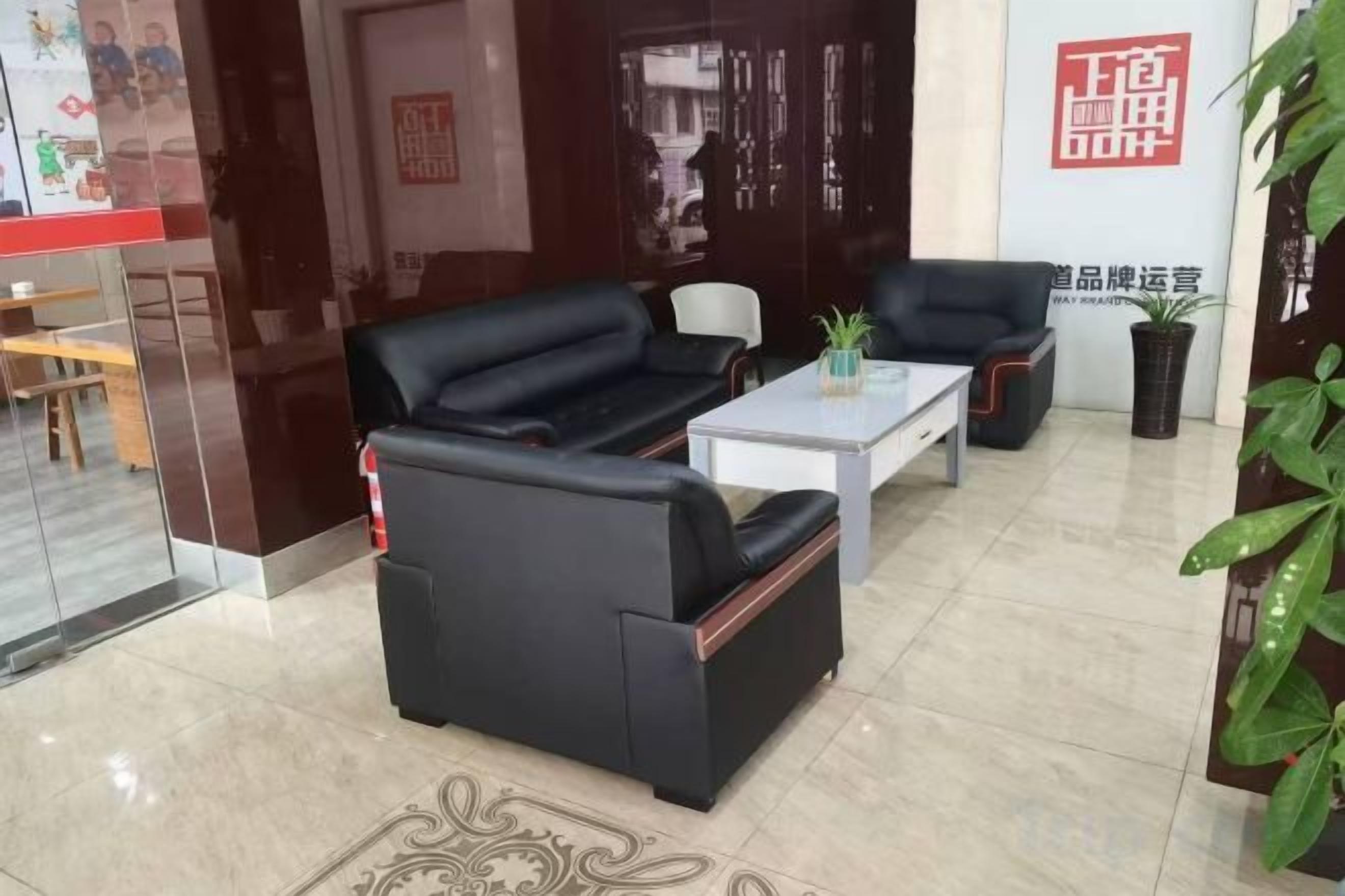 A hotel lobby with a black leather sofa, a white coffee table, and a black armchair. There is a potted plant on the coffee table and a red sign on the wall.