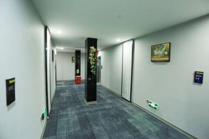 A hallway with a carpeted floor, a black pillar with a floral arrangement, and a painting on the wall.