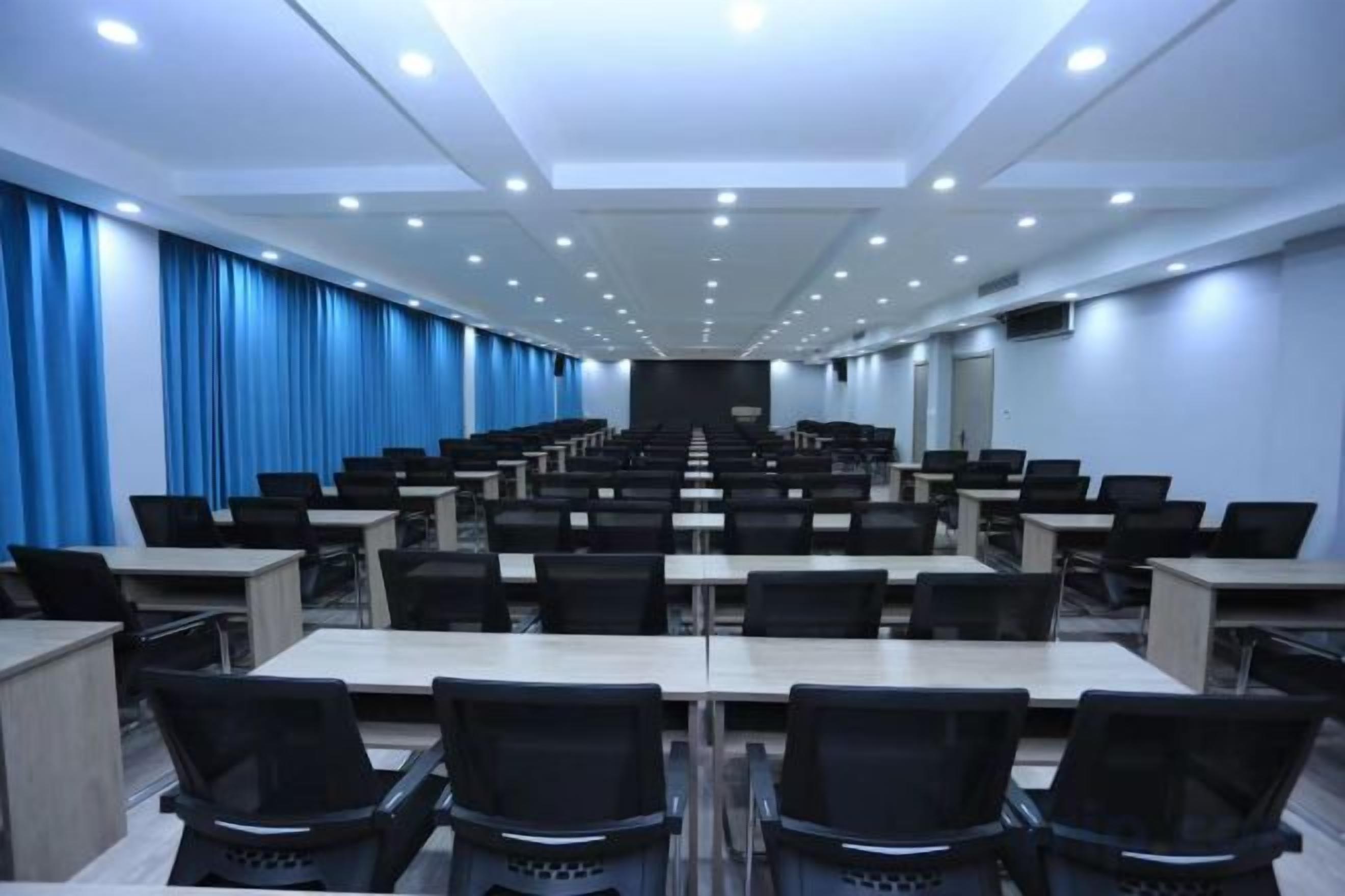 A conference room with rows of tables and chairs, a stage at the front, and blue curtains on the walls.