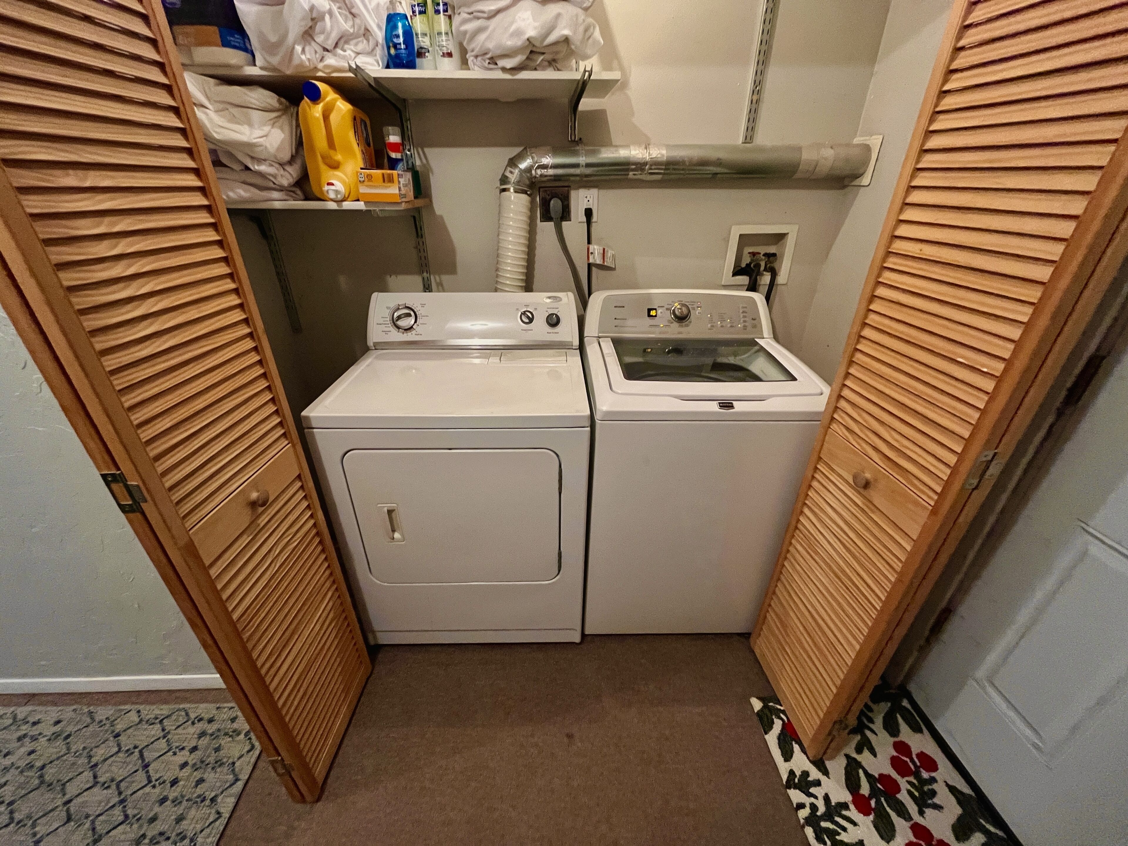 A compact laundry room with two white washing machines, a shelf with cleaning supplies, and a wooden door.