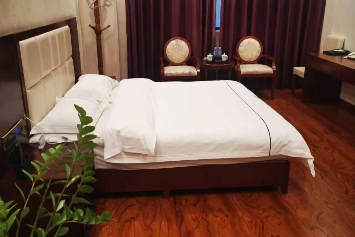 A neatly made bed with white linens, a wooden headboard, and a dark wooden floor. There are two wooden chairs with cushioned seats and a small round table with a lamp and a book.