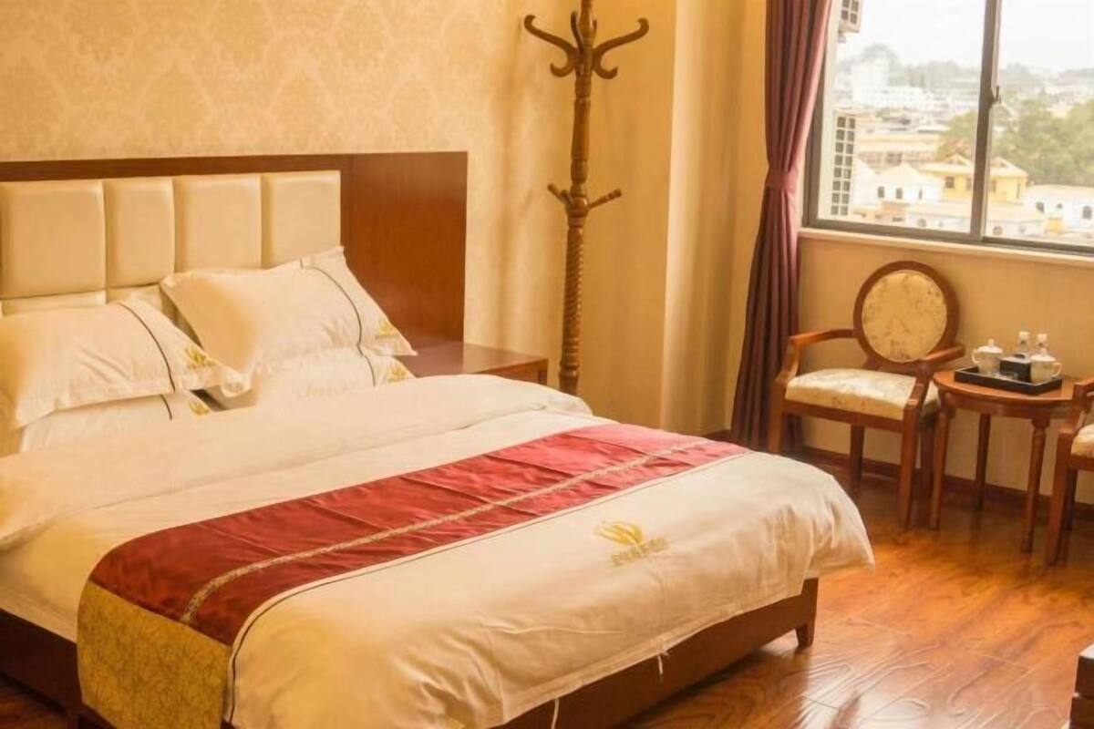 A hotel room with a bed, a chair, a small table with items, a coat rack, and a window with a view.
