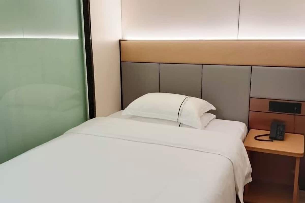 Una cama bien tendida con ropa blanca, una mesita de noche con un teléfono y una pared con un panel de luz.