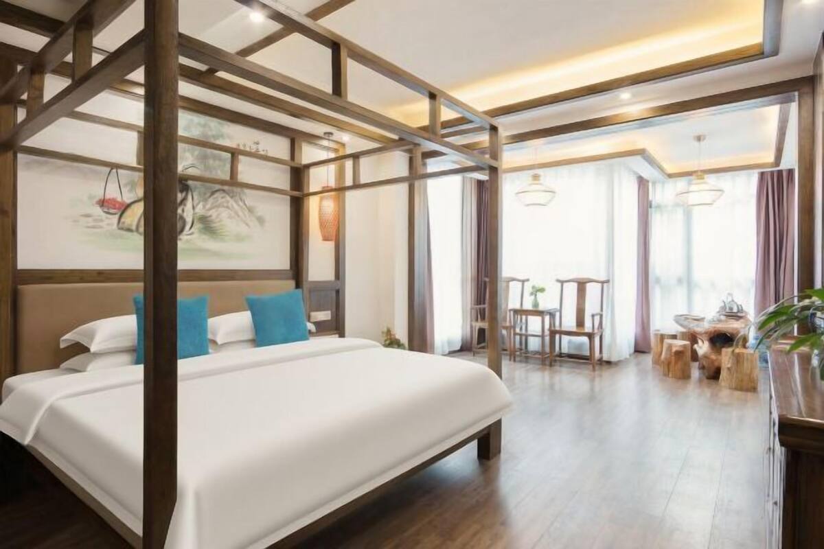 Une chambre d’hôtel avec un grand lit, des poutres apparentes, un coin repas avec une table et des chaises, et une vue par la fenêtre.