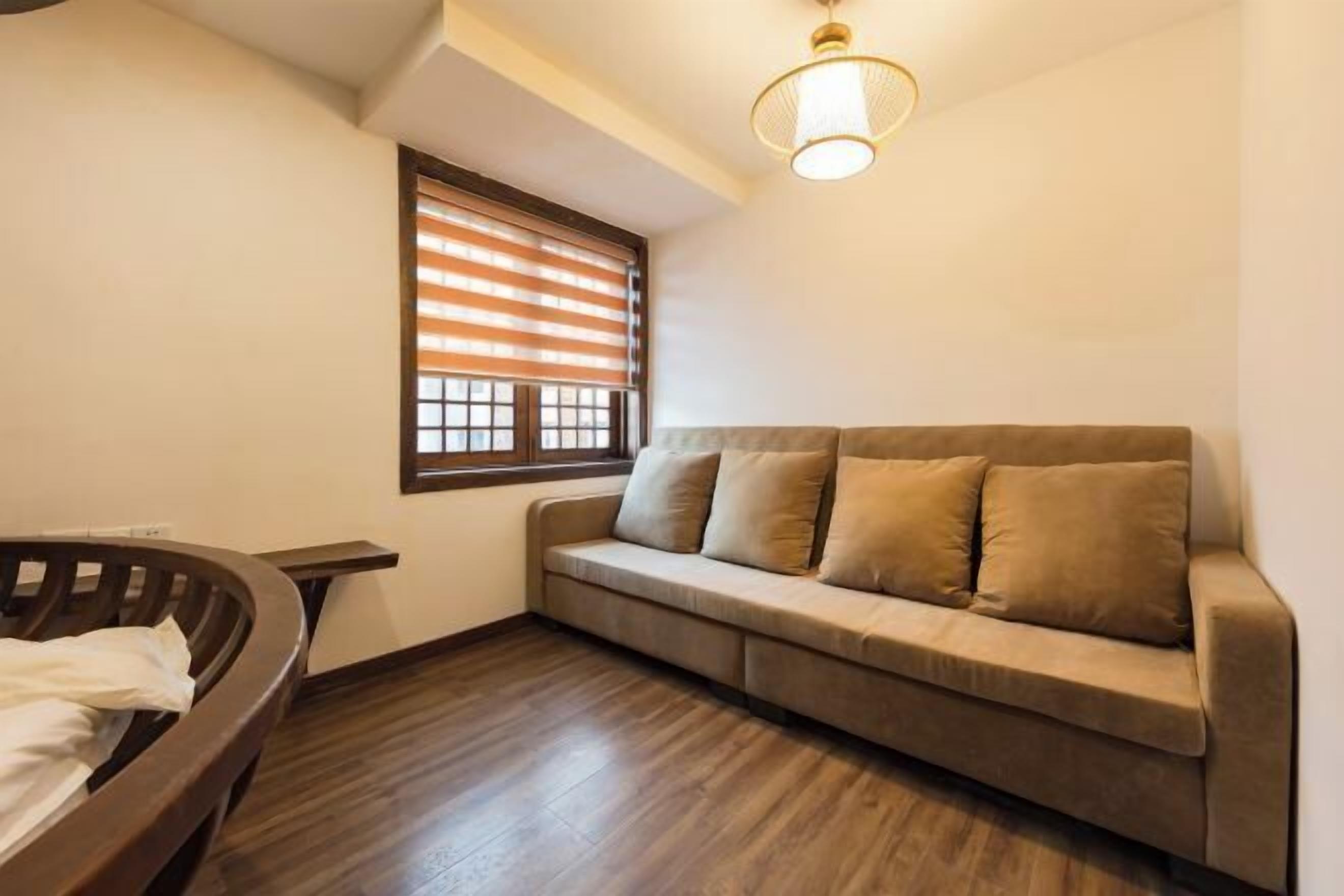 A room with a wooden floor, a beige sofa with cushions, a wooden bench, and a window with blinds.