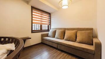 A room with a wooden floor, a beige sofa with cushions, a wooden bench, and a window with blinds.