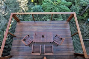 A wooden deck with a table and chairs, surrounded by a metal fence and lush greenery. - Aqua Vista - Pet friendly with yard and views (Merimbula)