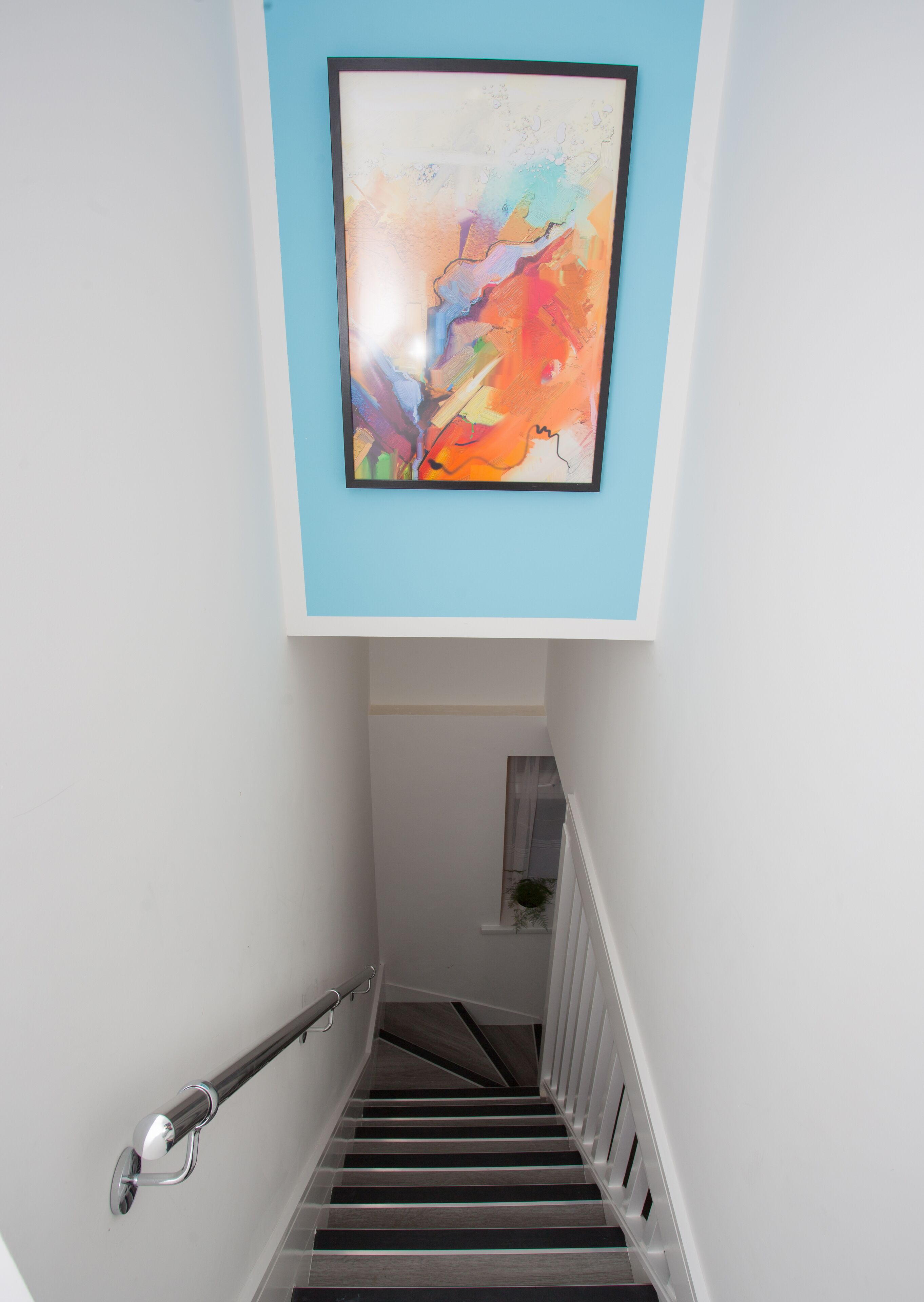 Un escalier muni d’une rampe, un tableau abstrait encadré accroché au mur et une petite plante en pot sur le palier.
