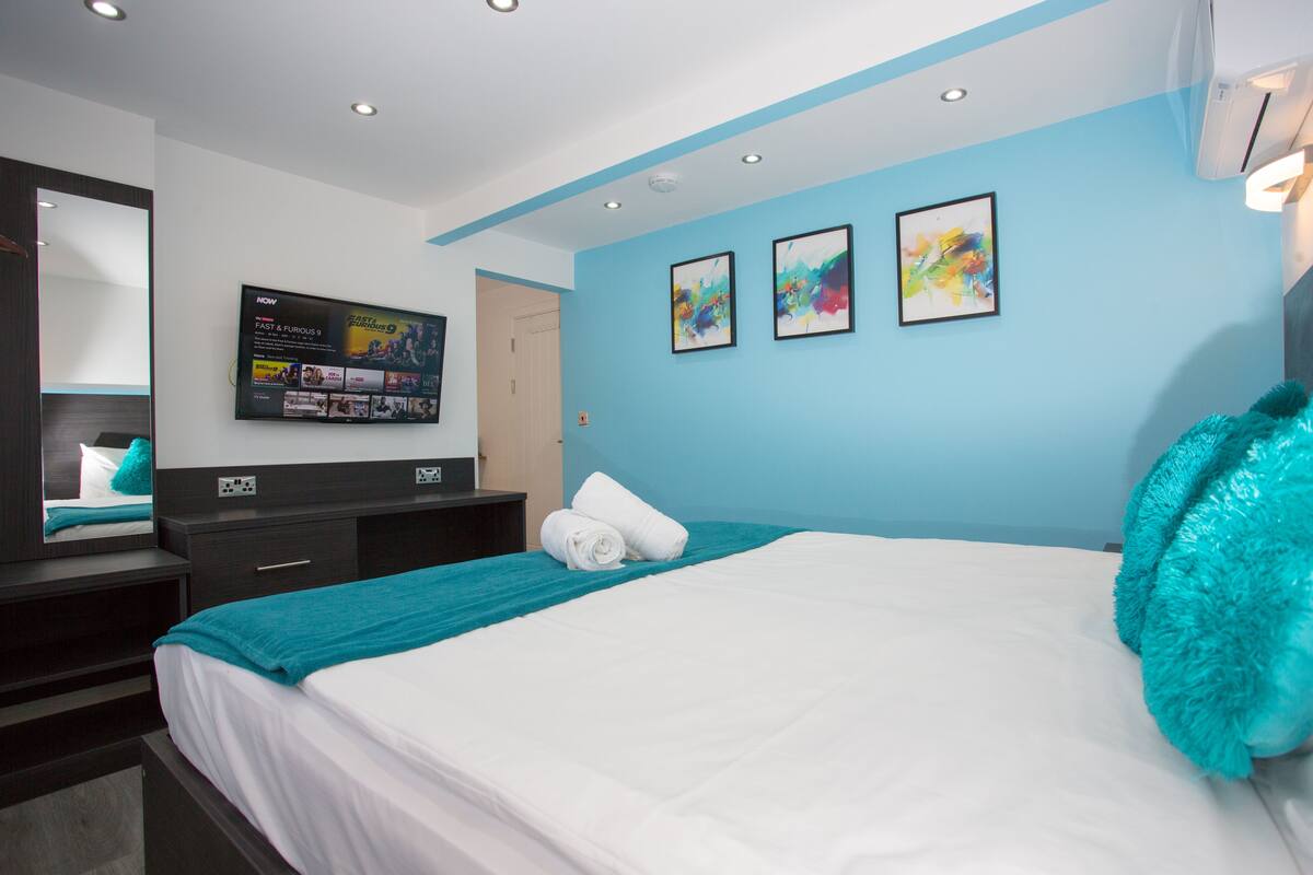 A modern hotel room with a large bed, a flat-screen TV mounted on the wall, and artwork on the blue walls.