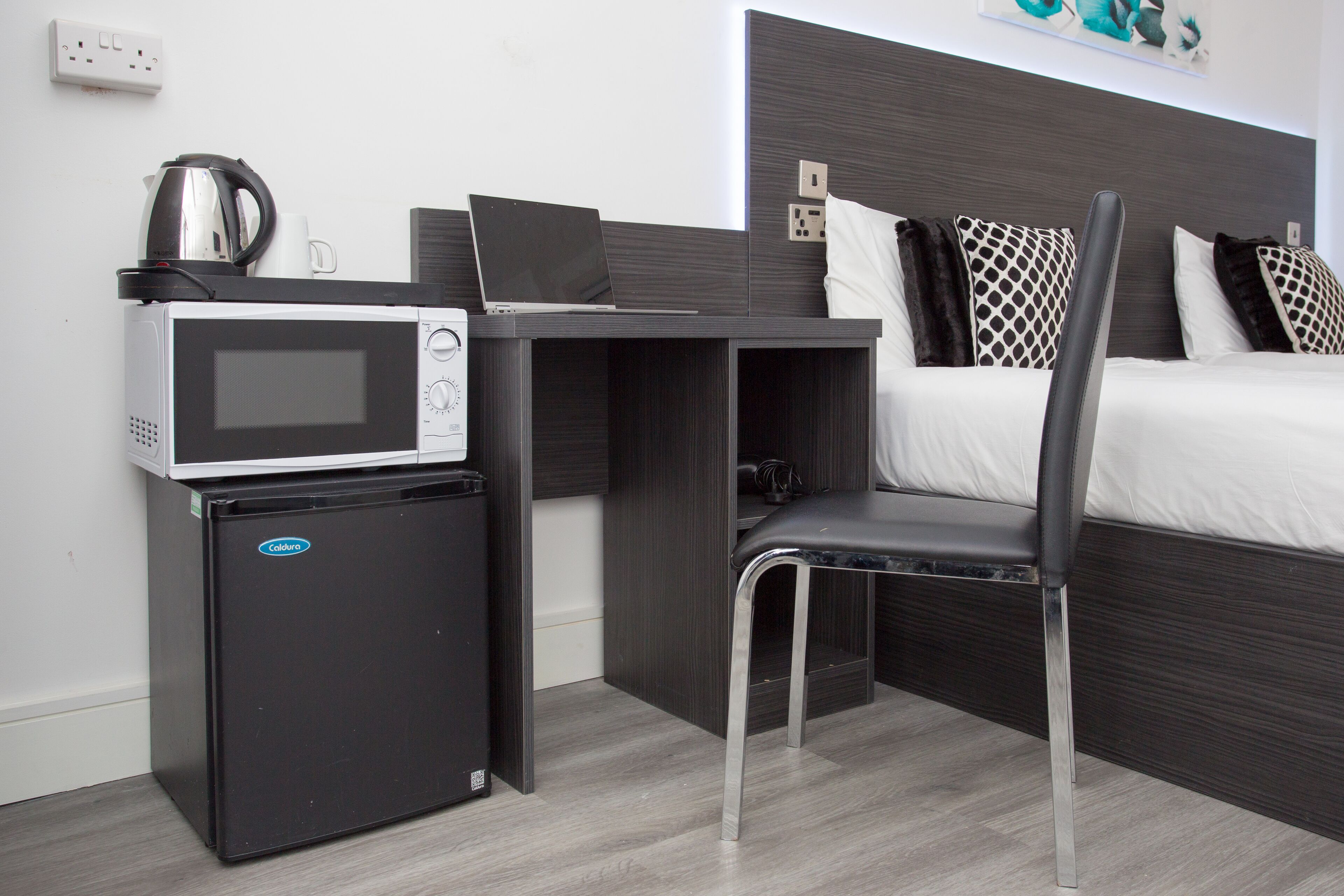 Una habitación de hotel compacta con una cama, un escritorio con una computadora portátil, un microondas y un refrigerador pequeño.