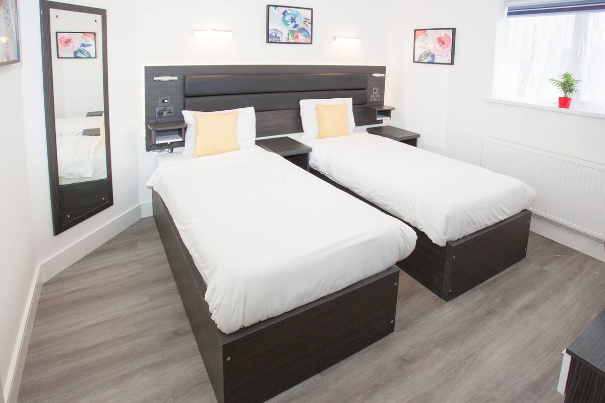 A hotel room with two single beds, a large mirror, bedside tables, and artwork on the walls.