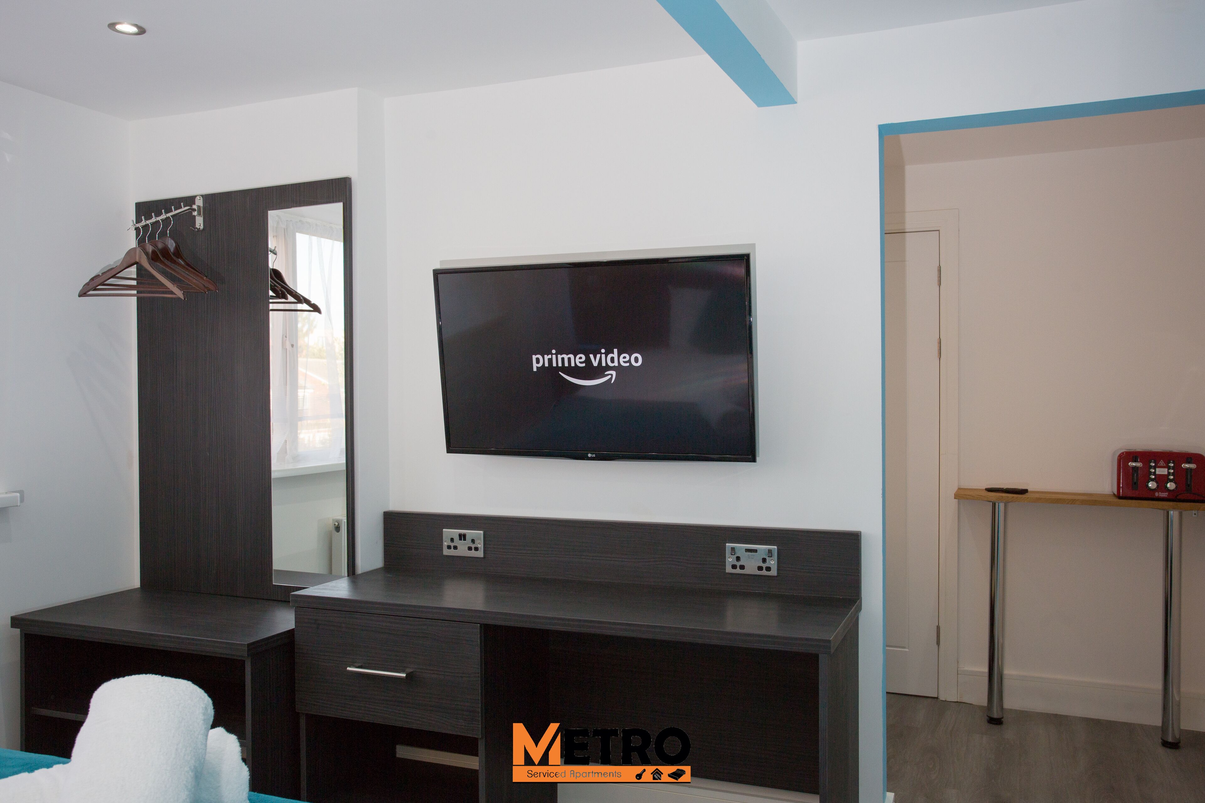 A modern bedroom with a large flat-screen TV mounted on the wall, a dark wooden dresser, a mirror, and a bedside table with a red radio.