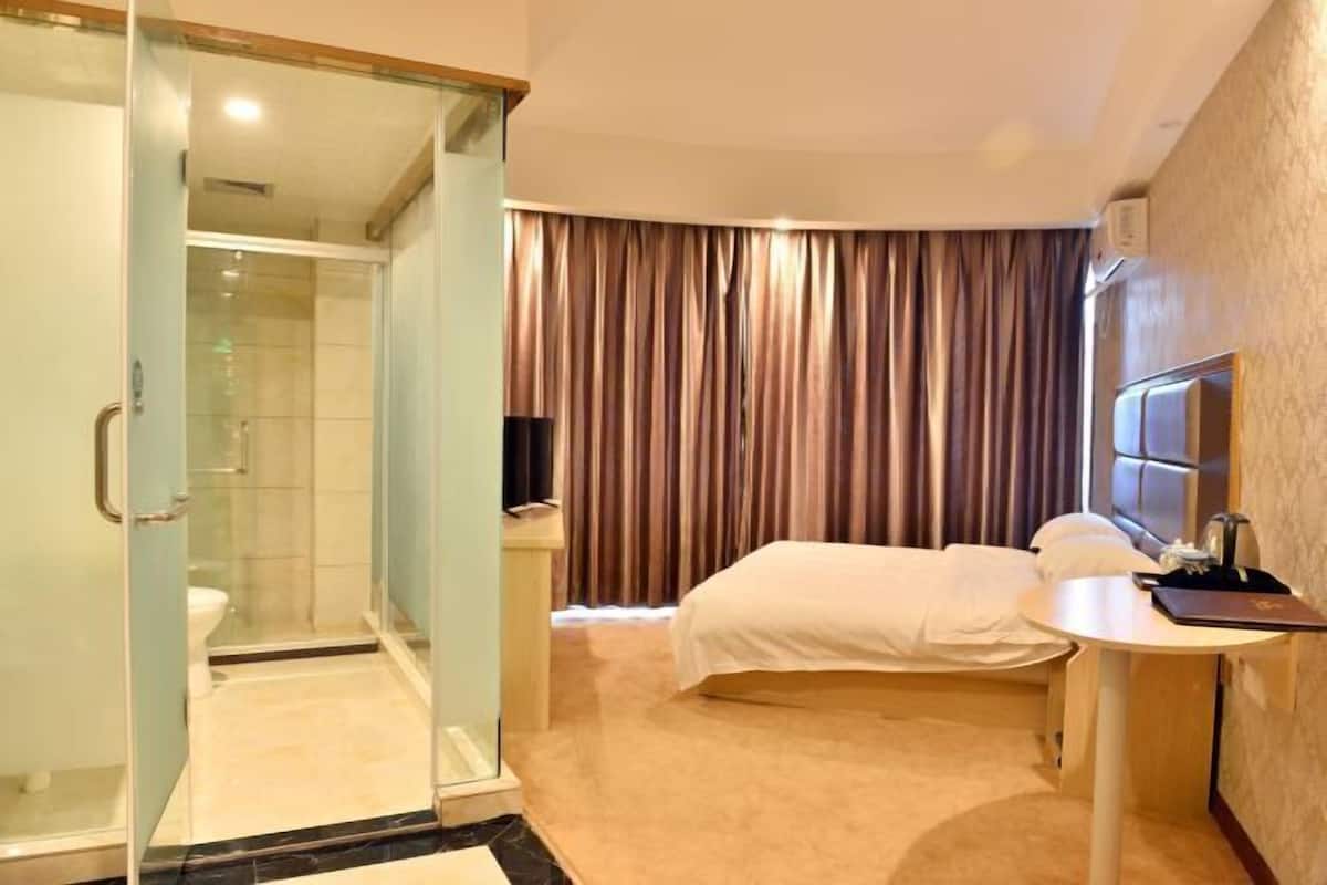 Een hotelkamer met een douchecabine van glas, een bed met een witte dekbedovertrek, een nachtkastje met een lamp en een groot raam met gordijnen.