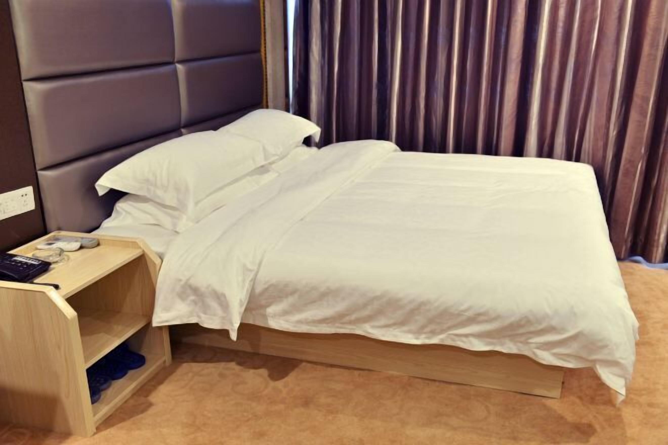 A neatly made bed with white bedding, a wooden nightstand with a phone and a book, and a headboard with a padded design.