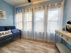 Una habitación con pared azul, piso de madera, sofá con almohada que dice 'BEACH' y ventana con cortinas blancas.