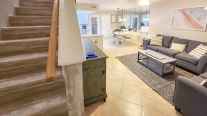 A spacious living area with a staircase, a sofa set, a coffee table, and a dining area in the background.
