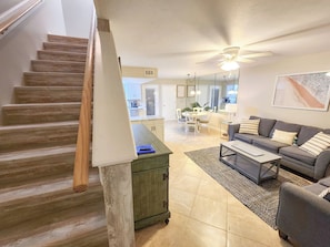 Una escalera conduce a una sala de estar con sofá y una mesa de centro, con una cocina al fondo y un ventilador de techo.