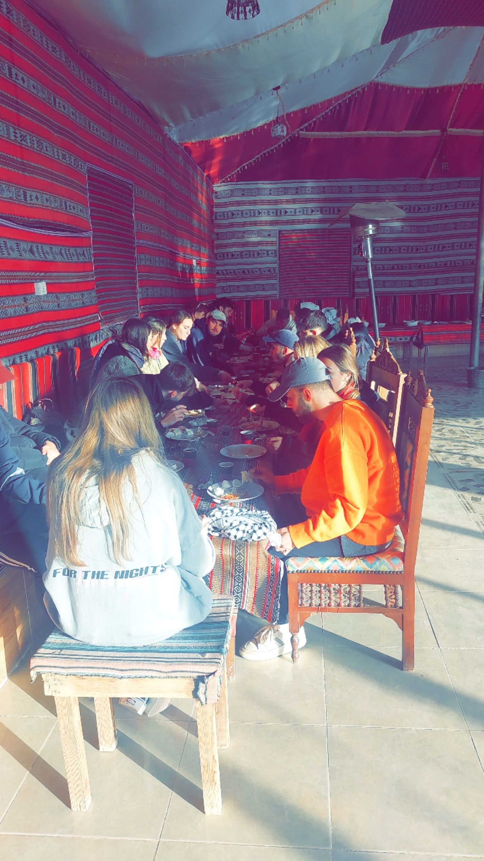 A group of people seated at a long table outdoors, under a tent-like structure.