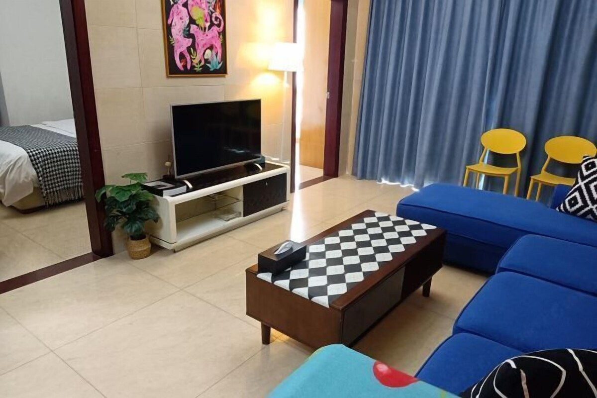A hotel room with a TV on a stand, a blue sofa, a checkered coffee table, and a bed with a checkered bedspread.