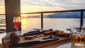 Una mesa con una copa de vino tinto, una botella y pan sobre una tabla de madera, con vistas a un cuerpo de agua y montañas al fondo.