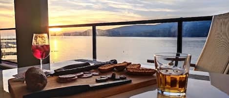 A table with a glass of red wine, a bottle, and some bread on a wooden board, overlooking a body of water with mountains in the background.