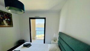 Habitación de hotel con cama, mesita de noche, lámpara y vista a la playa a través de una puerta abierta.
