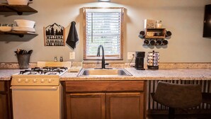 A kitchen with a stove, sink, and window with blinds. There are shelves with dishes and a sign that says "Welcome to our neck of the woods."