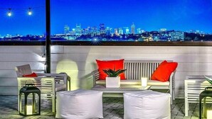 Una terraza en la azotea con mobiliario blanco, cojines rojos y vistas a un horizonte urbano nocturno.