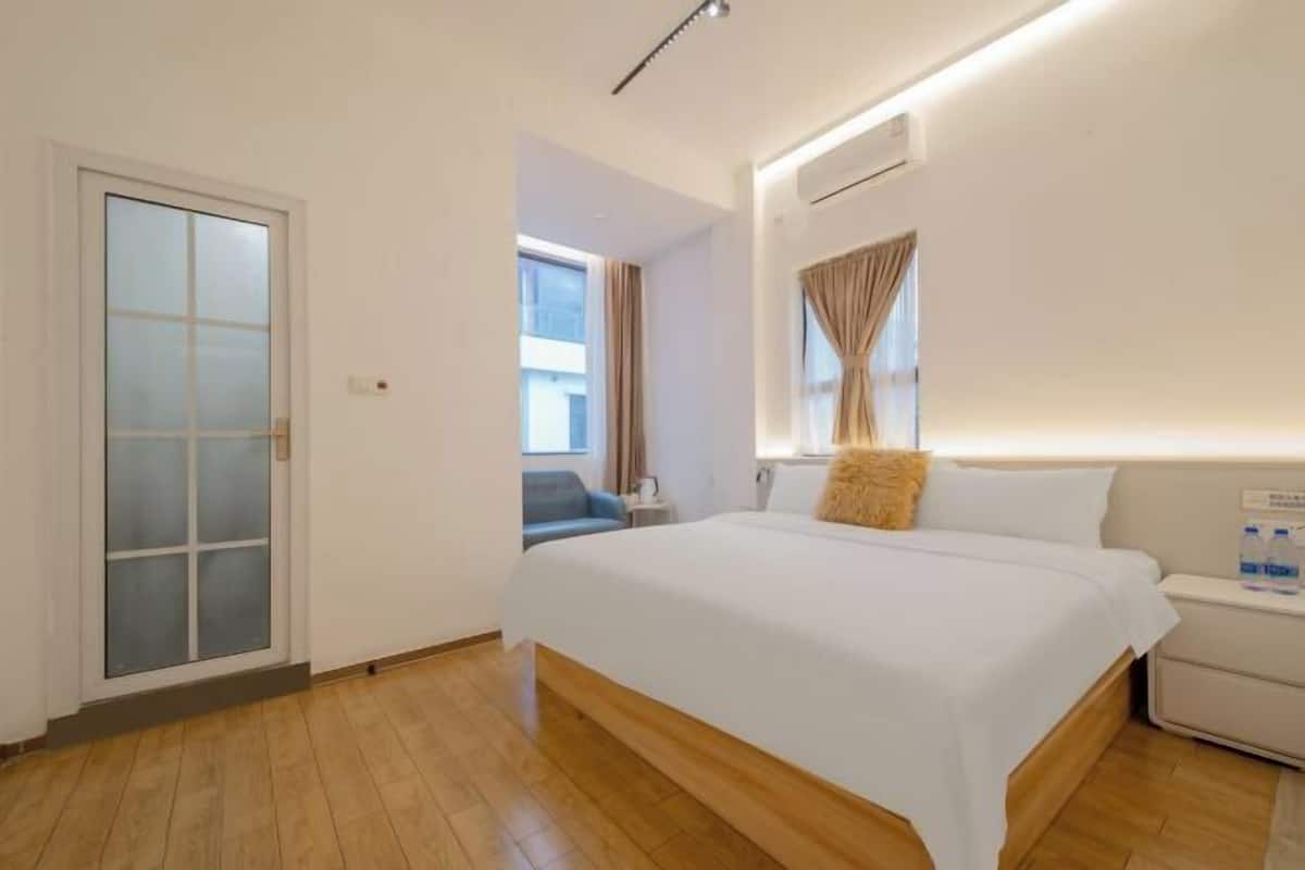 Una habitación de hotel con una cama grande, una mesita de noche, una ventana con cortinas y una puerta con paneles de vidrio esmerilado.