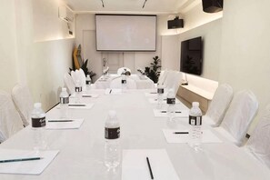 Une salle de conférence avec une longue table, des chaises blanches, des bouteilles d’eau, des stylos et un écran de projection.
