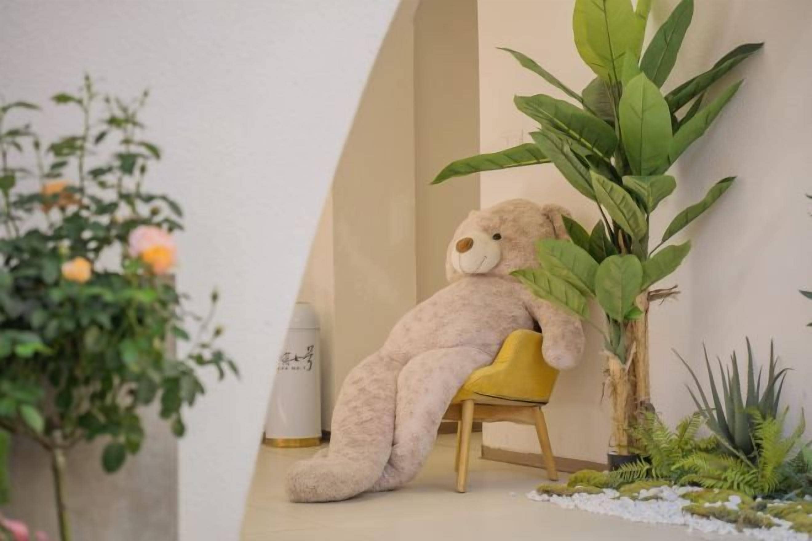 A teddy bear sitting on a yellow chair in a room with a white wall, a potted plant, and a white cylindrical object.