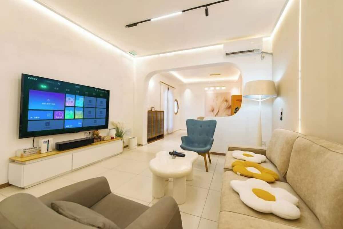 Una sala de estar moderna con una televisión de pantalla plana, un sofá con cojines en forma de huevo, un sillón azul y una mesa de centro blanca.
