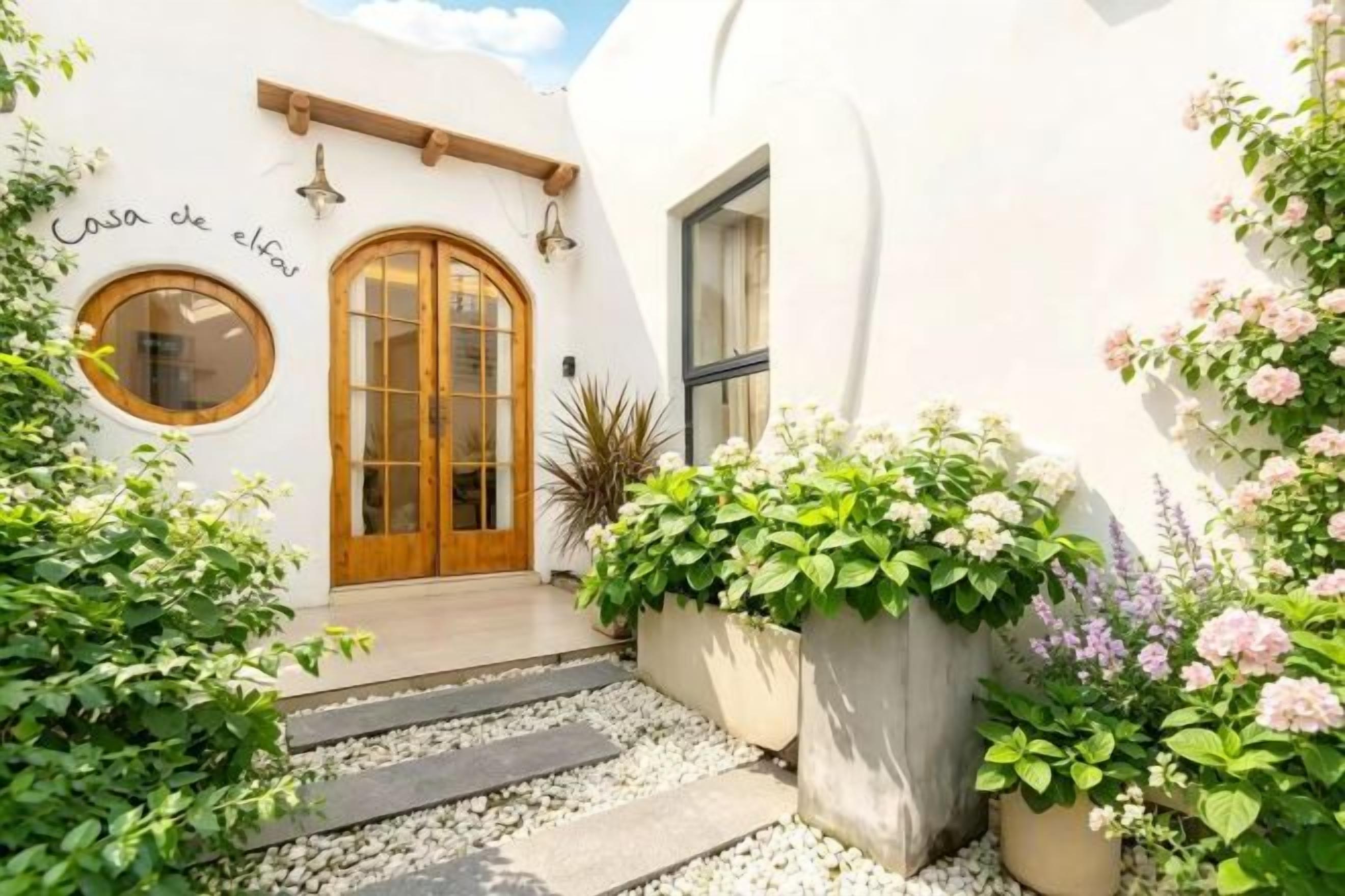 A white building with a wooden door and a circular window. There are steps leading to the entrance, and potted plants are placed around the area.