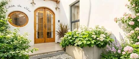 A white building with a wooden door and a circular window. There are steps leading to the entrance, and potted plants are placed around the area.