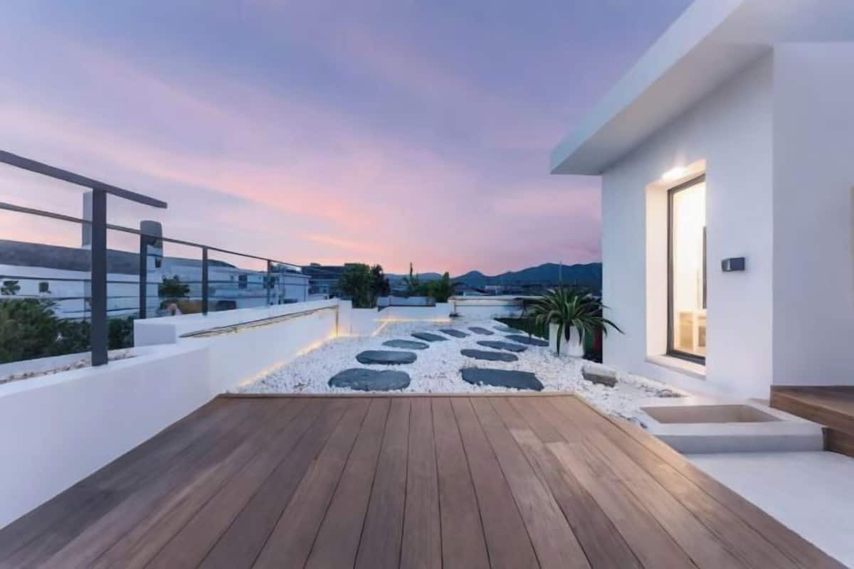 Una terraza moderna en la azotea con piso de madera, paredes blancas y vistas a montañas distantes bajo un cielo crepuscular.