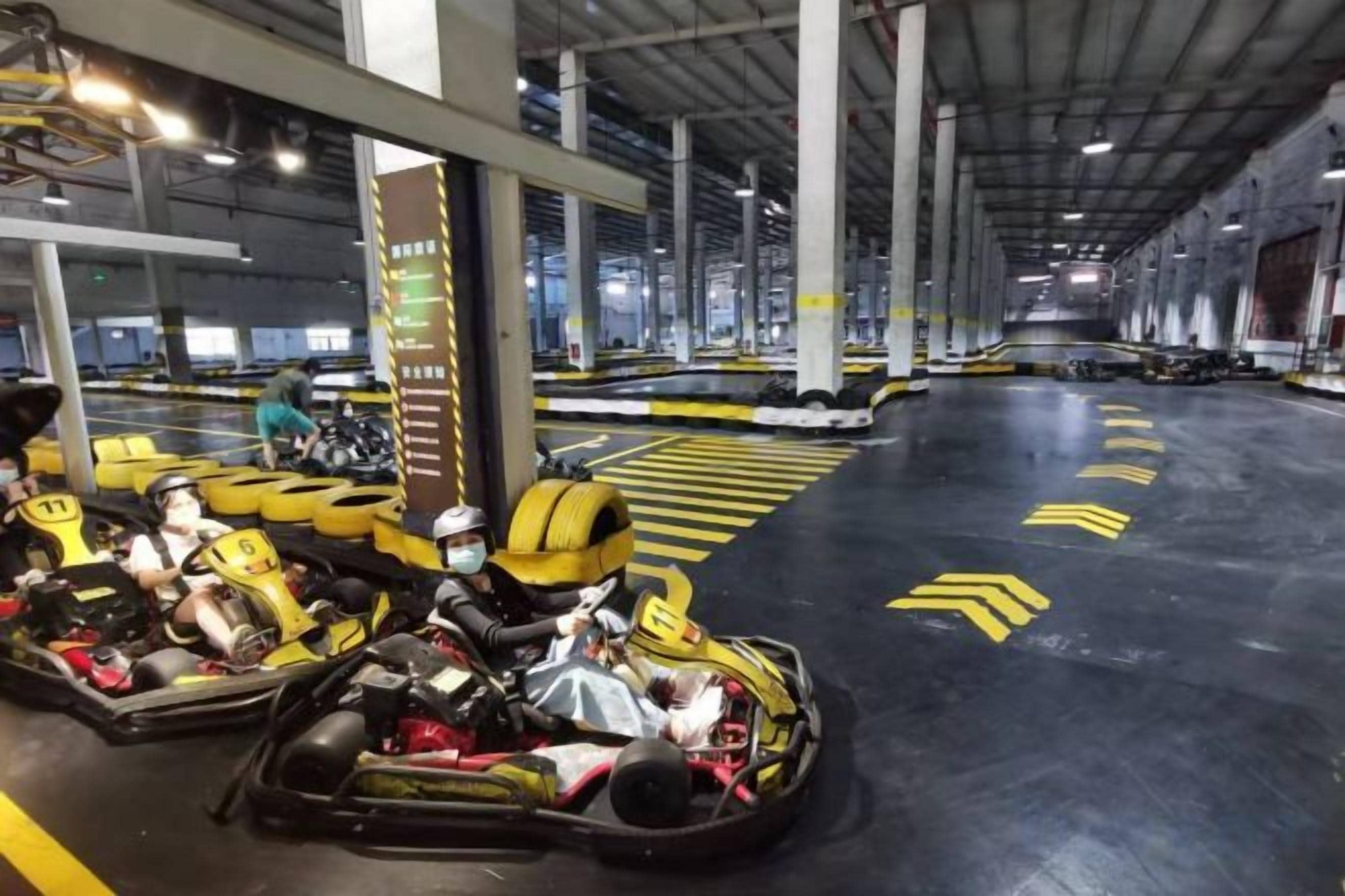 A go-kart track with multiple go-karts and riders, enclosed space with a high ceiling, and directional arrows on the floor.