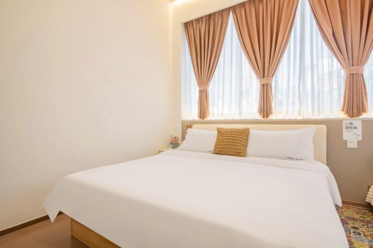 A neatly made bed with white linens and a single brown pillow, a window with beige curtains, and a small nightstand with a lamp.