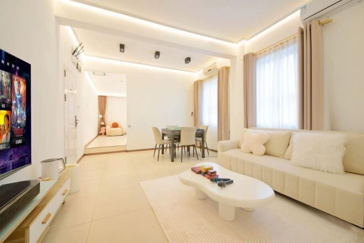 A modern living room with a sofa, coffee table, and TV. There is a dining area with a table and chairs.