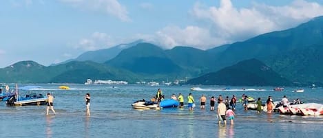 A beach with boats and people, mountains in the background, and clear skies.