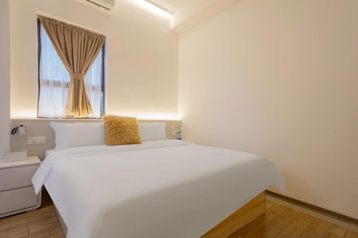A hotel room with a large bed, a nightstand, a window with curtains, and a wall-mounted air conditioning unit.