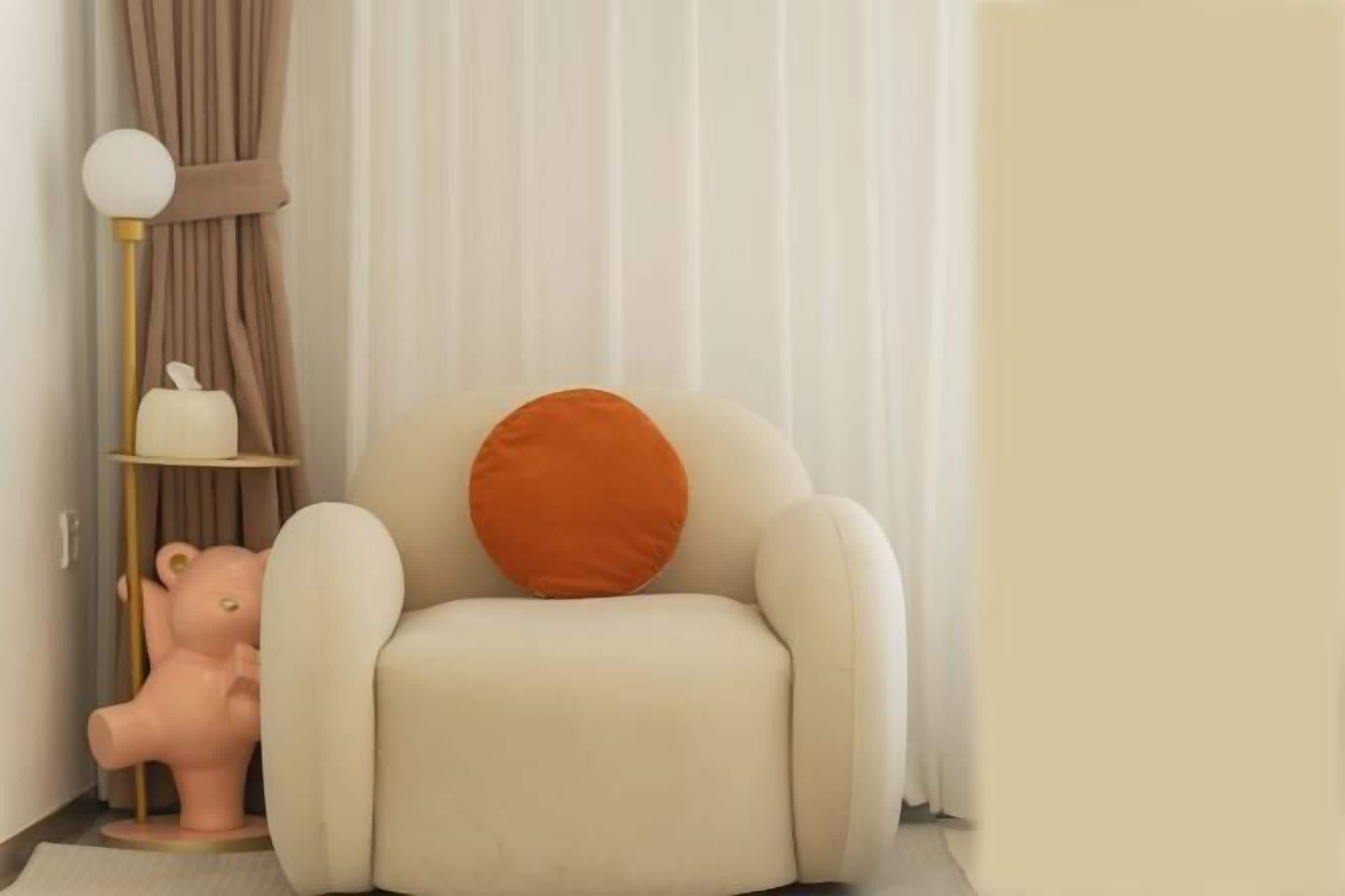 A room with a beige armchair, a small orange pillow, a pink bear-shaped object, and a floor lamp with a white globe.