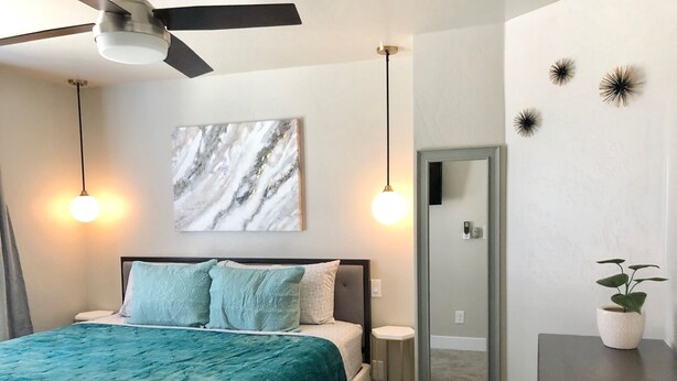 Ein Schlafzimmer mit Bett, Deckenventilator, Marmorbild, Nachttischlampen, einem kleinen Nachttisch mit Pflanze und einem Spiegel.