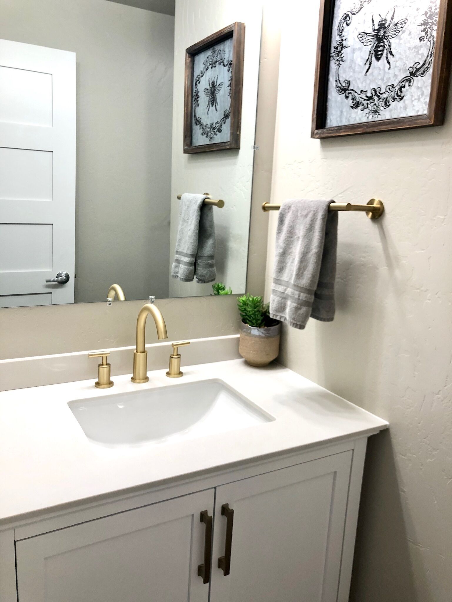 Une salle de bain avec un lavabo blanc, des robinets dorés et un miroir au-dessus. Deux serviettes sont suspendues à des porte-serviettes et une petite plante est posée sur le comptoir du lavabo.