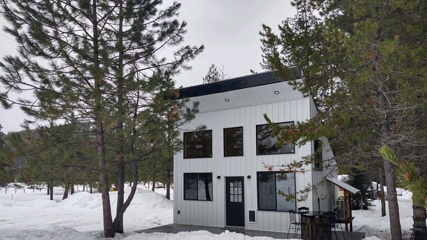 Una cabaña de dos pisos con fachada blanca, rodeada de nieve y pinos.