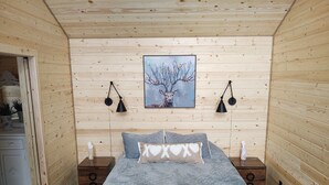 Un dormitorio acogedor con un revestimiento de madera, una cama con ropa de cama gris, dos lámparas de noche y un cuadro de un árbol con un ciervo.