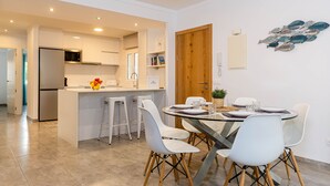 A modern kitchen with white cabinets, a built-in microwave, and a refrigerator. There is a dining area with a glass table and white chairs. - YourHouse Can Cortana (Pollensa)