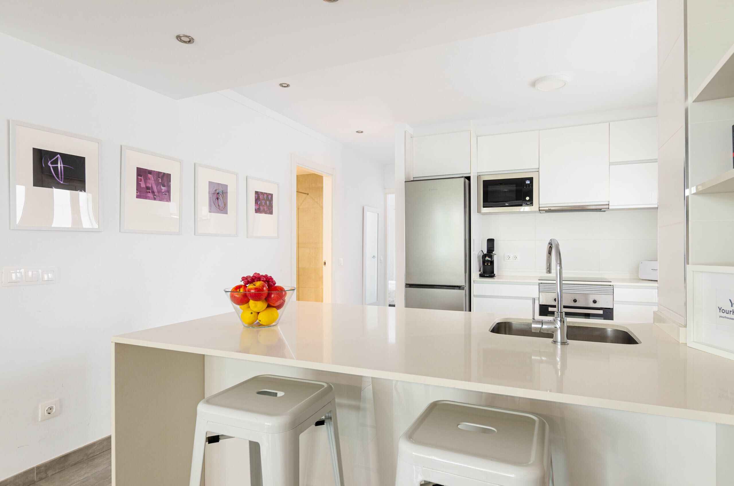 Een moderne keuken met witte kasten, een roestvrijstalen koelkast, een magnetron en een spoelbak. Er hangen prestaties aan de muur en op het aanrecht staat een fruitschaal.