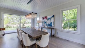 Enjoy meals in this dining area with a large table and six chairs, surrounded by a high ceiling and large windows offering views of the surrounding greenery.